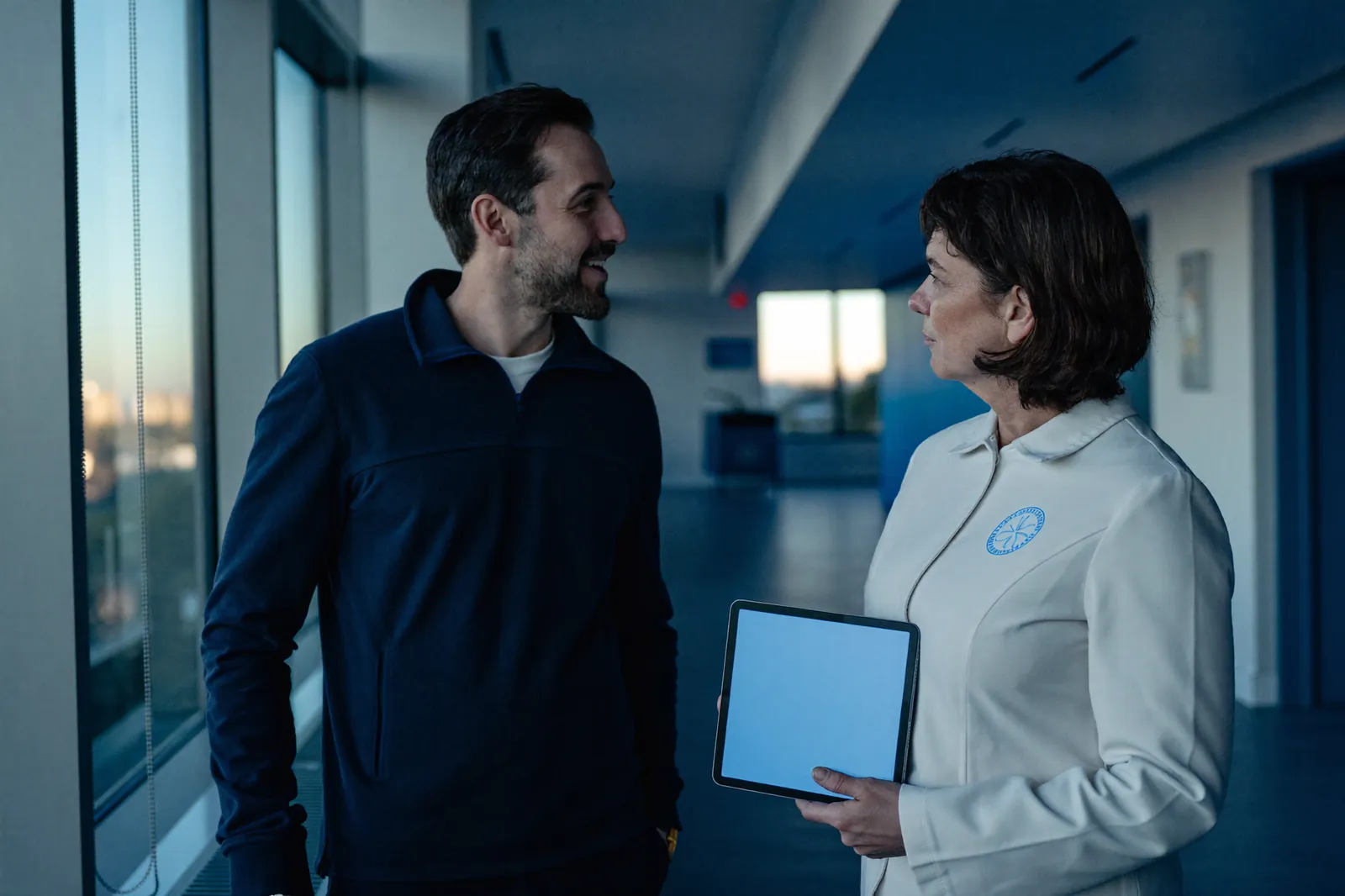 Two clinicians talk beside a tall window in a clinic corridor, one holding a tablet.