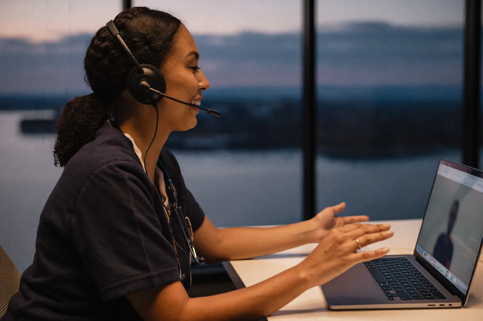 A nurse practitioner wearing a headset speaks with a patient on a telehealth video call from a clinic office at dusk.