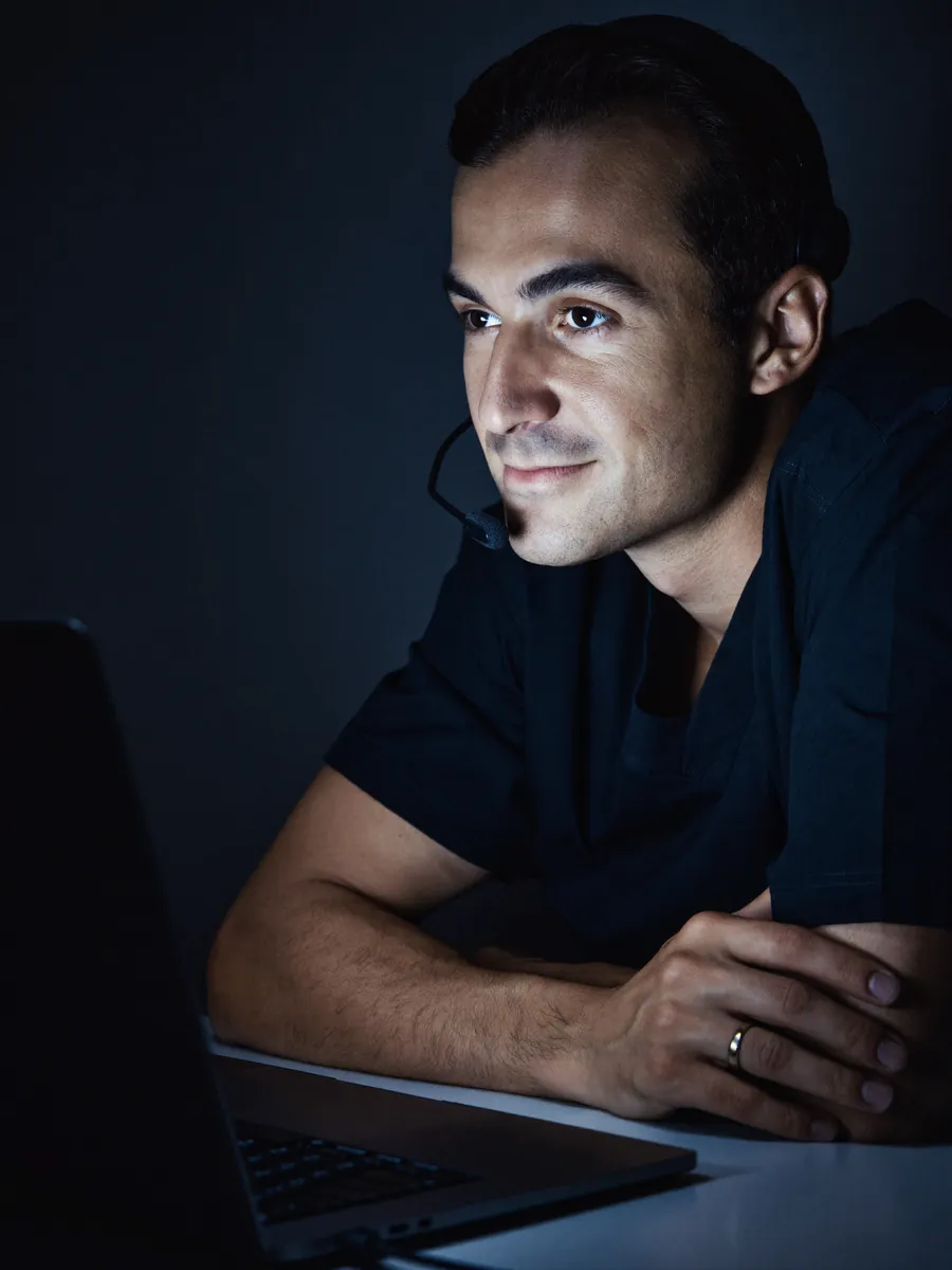 A physician assistant wearing a headset on a video visit, lit by his laptop screen at night.