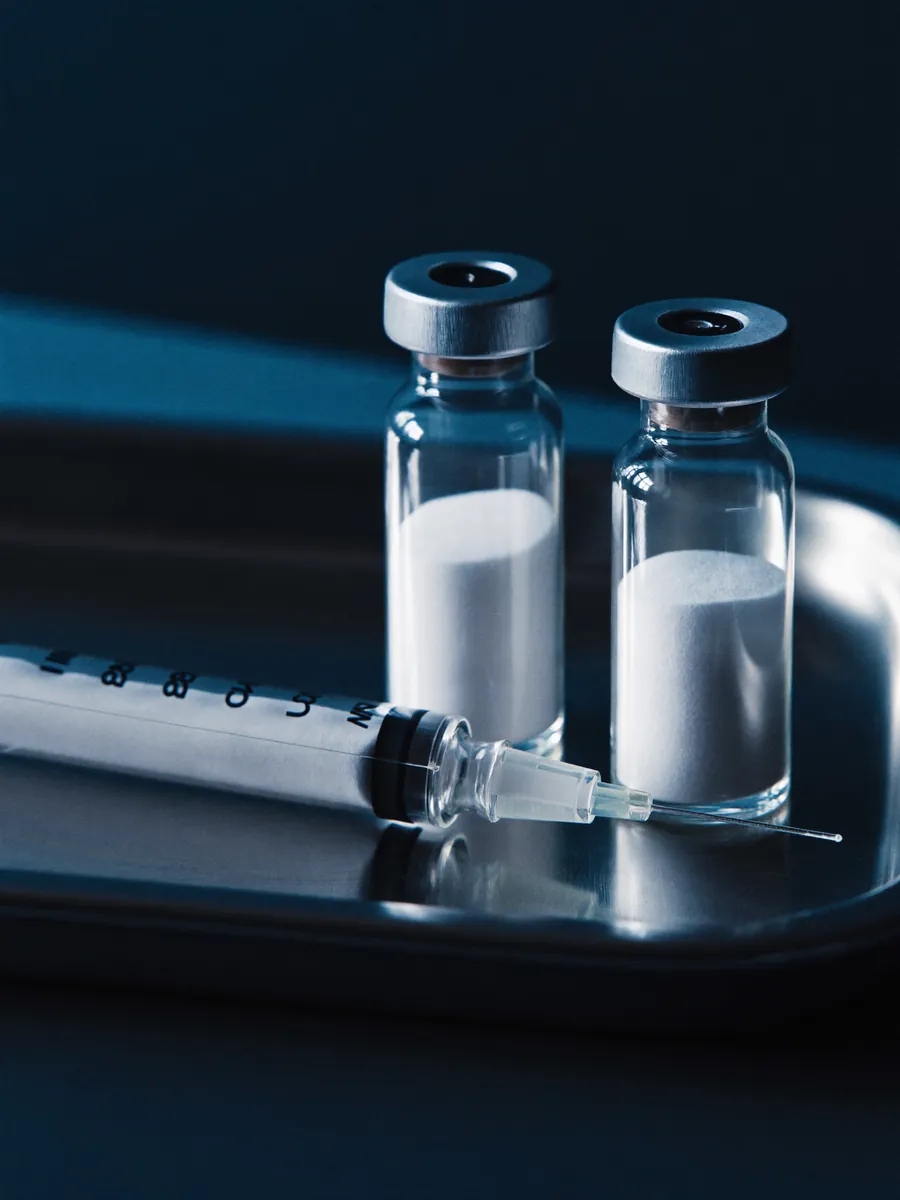Two small glass vials of lyophilized peptide beside a syringe on a steel tray.