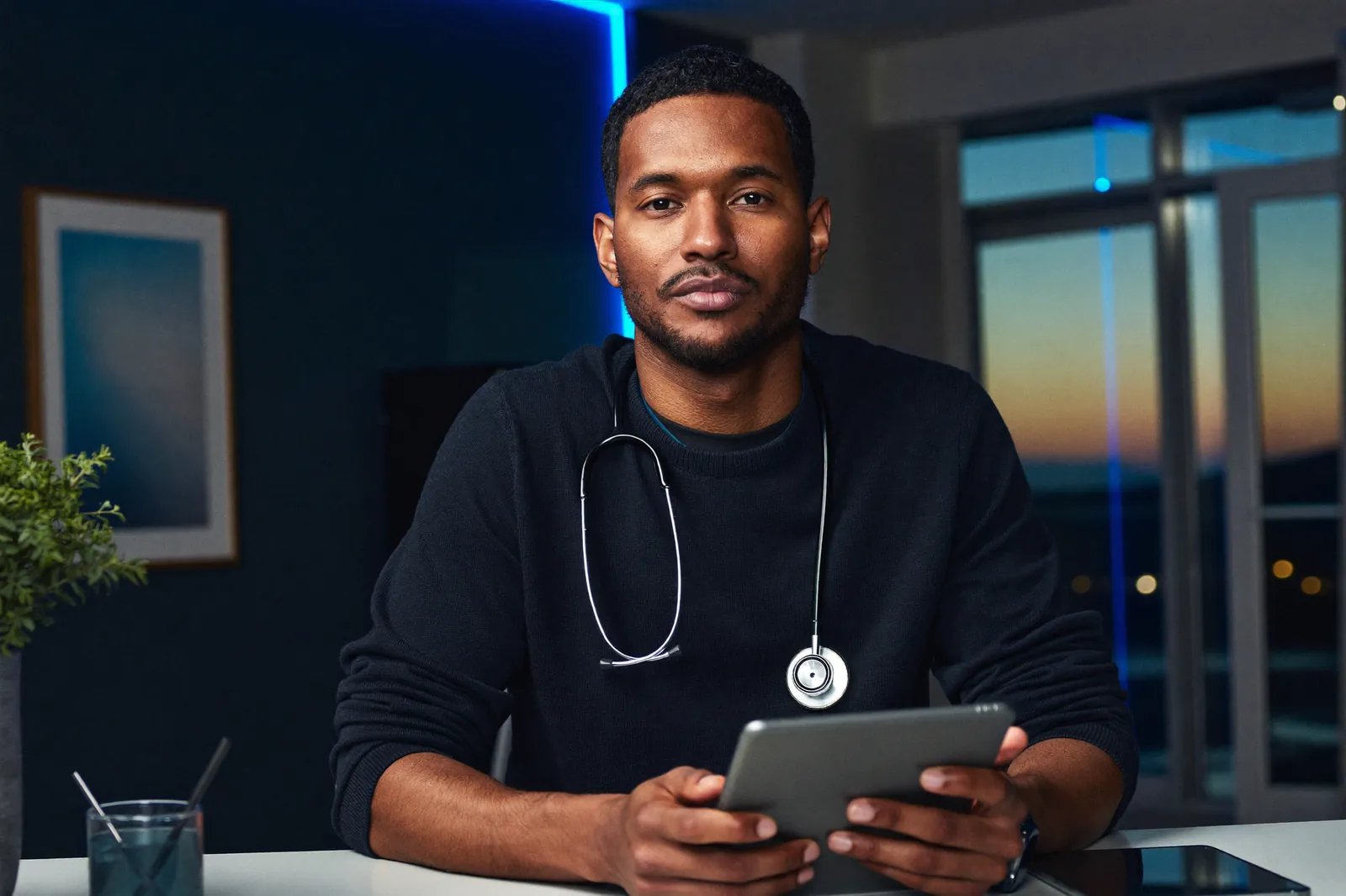 A physician with a stethoscope holds a tablet and looks directly at the camera in a clinic at dusk.